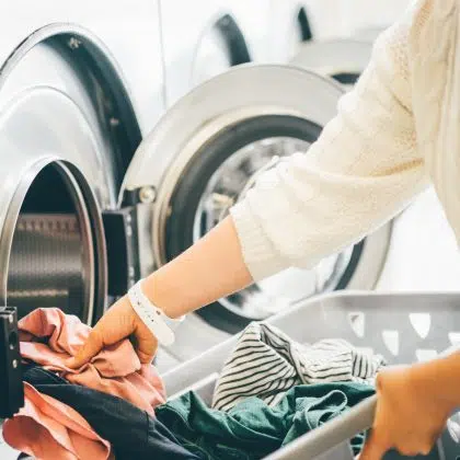 Interior of small laundromat in daylight. Close-up female holding basket. Girl loading dirty clothes inside drum and closing door. Self-service concept